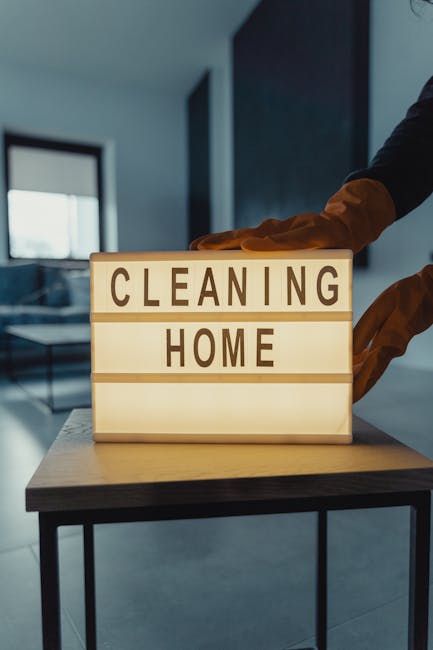 A woman wearing an orange apron and gray t-shirt is performing surface cleaning in a modern kitchen, using a mop on the light-colored floor. The kitchen features sleek white walls, a built-in microwav